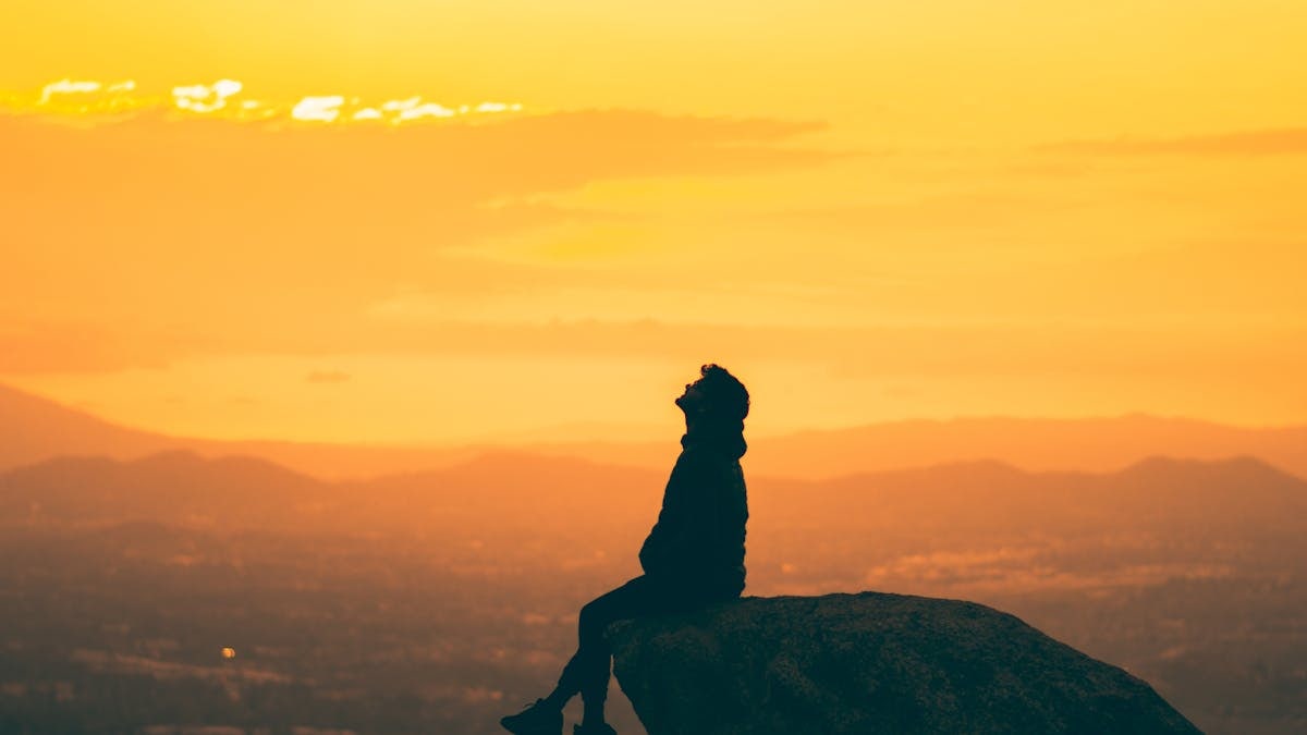 A contemplative silhouette sits on a rock during a serene sunset, evoking tranquility.